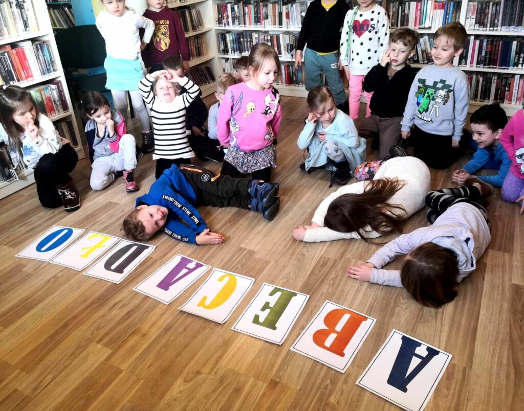 na zdjęciu widać dzieci w sali bibliotecznej a przed nimi na podłodze napis abecadło