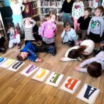 na zdjęciu widać dzieci w sali bibliotecznej a przed nimi na podłodze napis abecadło