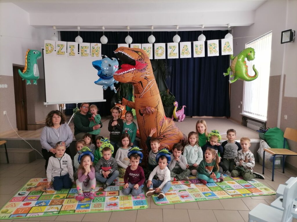 na zdjęciu widoczna jest grupa przedszkolaków (10 dziewczynek i 9 chłopców) wraz z 3 opiekunkami, siedzących na kolorowych matach i pozujących do wspólnego zdjęcia z dmuchanym dinozaurem. Nad dziećmi jest rozwieszony napis „dzień dinozaura” oraz kolorowe dmuchane dinozaury w tle pozostałe dekoracje nawiązujące do obchodzonego „dnia dinoaura”.