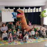 na zdjęciu widoczna jest grupa przedszkolaków (10 dziewczynek i 9 chłopców) wraz z 3 opiekunkami, siedzących na kolorowych matach i pozujących do wspólnego zdjęcia z dmuchanym dinozaurem. Nad dziećmi jest rozwieszony napis „dzień dinozaura” oraz kolorowe dmuchane dinozaury w tle pozostałe dekoracje nawiązujące do obchodzonego „dnia dinoaura”.