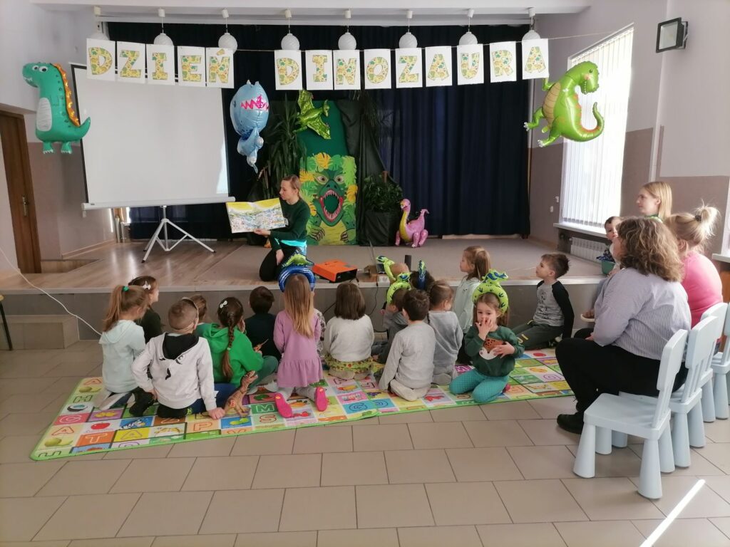 na zdjęciu widoczny jest bibliotekarz czytający bajkę dzieciom, które siedzą na kolorowych matach. Wokół dekoracje nawiązujące do obchodzonego „dnia dinozaura”.