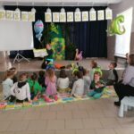 na zdjęciu widoczny jest bibliotekarz czytający bajkę dzieciom, które siedzą na kolorowych matach. Wokół dekoracje nawiązujące do obchodzonego „dnia dinozaura”.