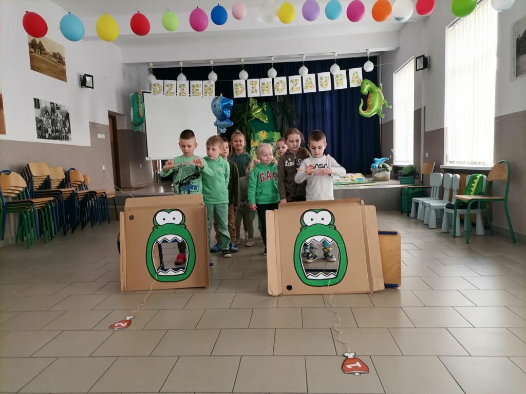 Na zdjęciu widoczne są dzieci ustawione w dwóch rzędach podczas zabawy w karmienie dinozaura. Polegającej na zawijaniu kości na patyk tak aby trafiła ona do paszczy dinozaura. W tle dekoracje nawiązujące do dnia dinozaura.
