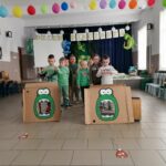Na zdjęciu widoczne są dzieci ustawione w dwóch rzędach podczas zabawy w karmienie dinozaura. Polegającej na zawijaniu kości na patyk tak aby trafiła ona do paszczy dinozaura. W tle dekoracje nawiązujące do dnia dinozaura.