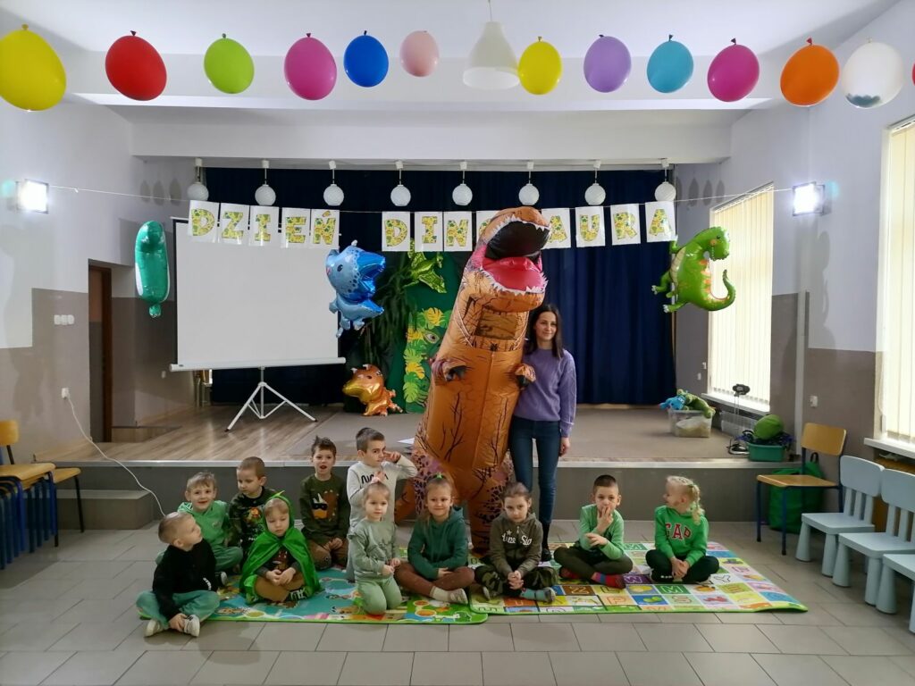 – Na zdjęciu widoczna jest grupa przedszkolaków (7 chłopców i 4 dziewczynki) pozujących do wspólnego zdjęcia z panią wychowawczynią i dmuchanym dinozaurem. Dzieci siedzą na kolorowych matach. Nad nimi jest rozwieszony napis „dzień dinozaura” oraz balony w kształcie dinozaurów. W tle dekoracje nawiązujące do obchodzonego dnia dinozaura.