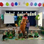 – Na zdjęciu widoczna jest grupa przedszkolaków (7 chłopców i 4 dziewczynki) pozujących do wspólnego zdjęcia z panią wychowawczynią i dmuchanym dinozaurem. Dzieci siedzą na kolorowych matach. Nad nimi jest rozwieszony napis „dzień dinozaura” oraz balony w kształcie dinozaurów. W tle dekoracje nawiązujące do obchodzonego dnia dinozaura.