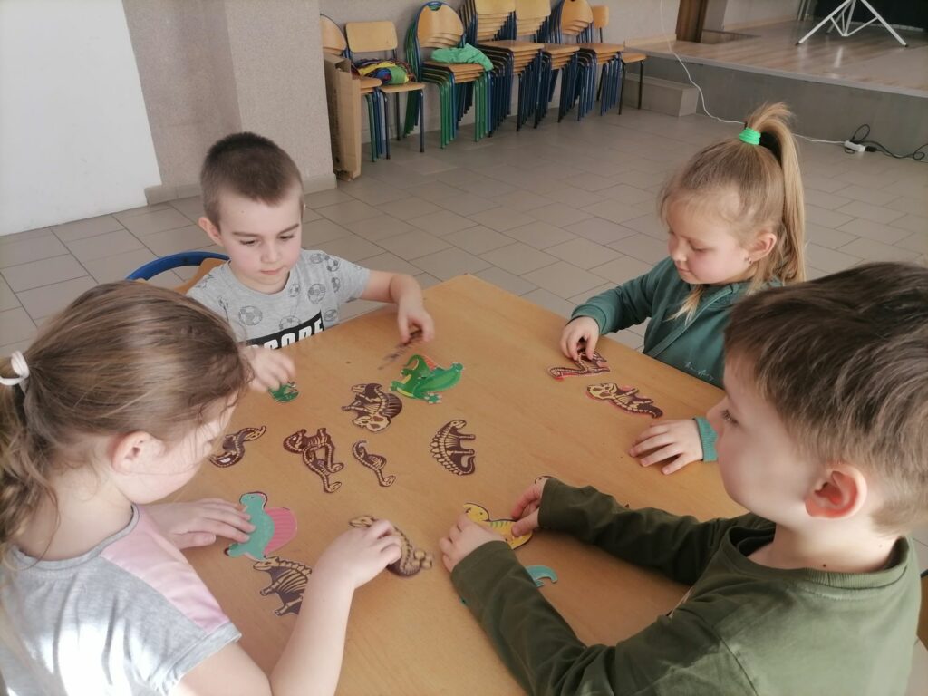 Na zdjęciu widocznych jest dwóch chłopców i dwie dziewczynki siedzących przy stoliku i dopasowujących szkielety dinozaurów do ich sylwetek.