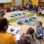 na zdjęciu widać dzieci siedzące w sali przedszkolnej na kolorowym dywanie i słuchające czytającej bibliotekarki.