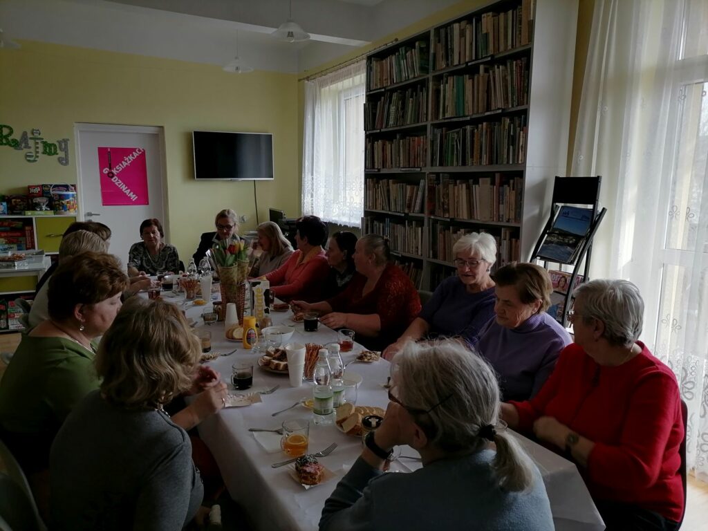 – Na zdjęciu widoczna jest grupa 18 pań, które spotkały się w bibliotece z okazji dnia kobiet. Panie siedzą przy stołach, na których jest drobny poczęstunek oraz wazon z kwiatami. W tle regały z książkami.