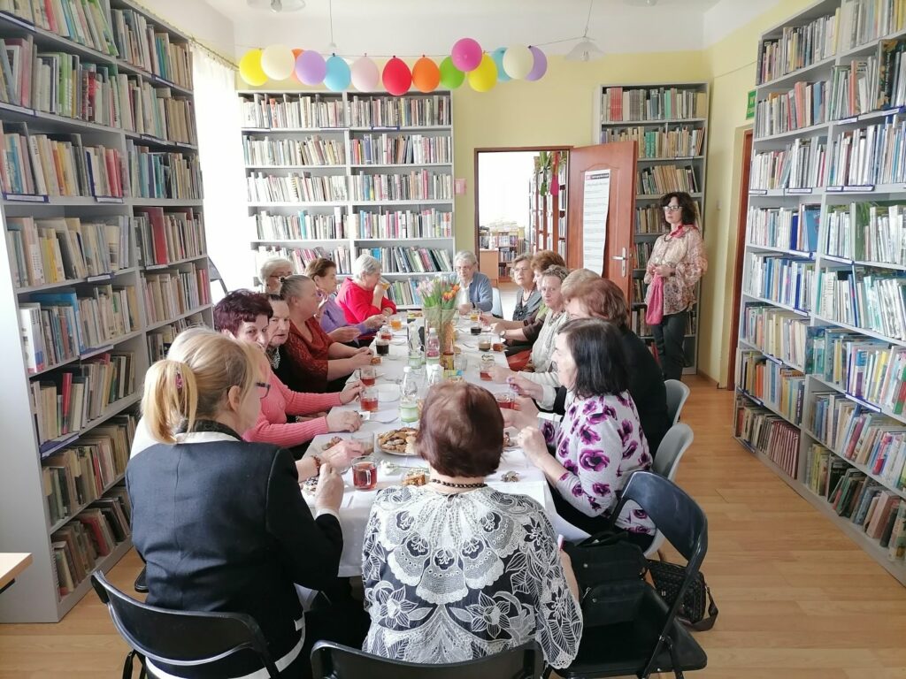 – Na zdjęciu widoczna jest grupa 18 pań, które spotkały się w bibliotece z okazji dnia kobiet. Panie siedzą przy stołach, na których jest drobny poczęstunek oraz wazon z kwiatami. W tle regały z książkami.