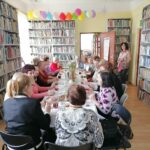 – Na zdjęciu widoczna jest grupa 18 pań, które spotkały się w bibliotece z okazji dnia kobiet. Panie siedzą przy stołach, na których jest drobny poczęstunek oraz wazon z kwiatami. W tle regały z książkami.