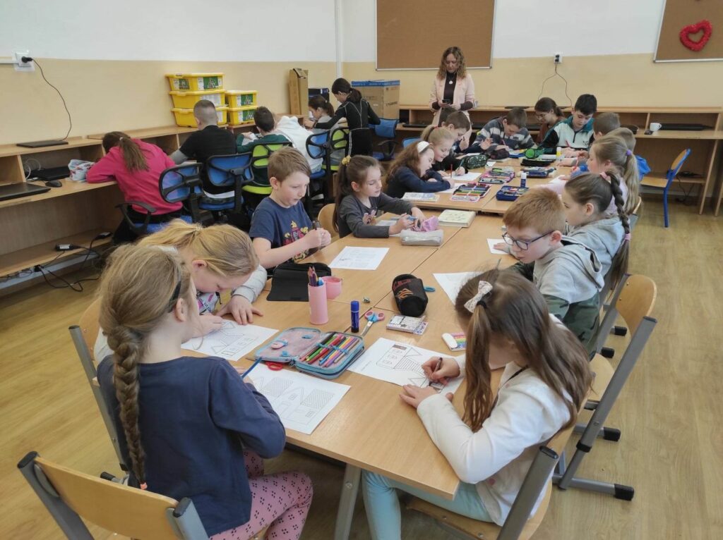 na zdjęciu widać salę szkolną, przy dużym stole siedzą dzieci, wykonują pracę plastyczną