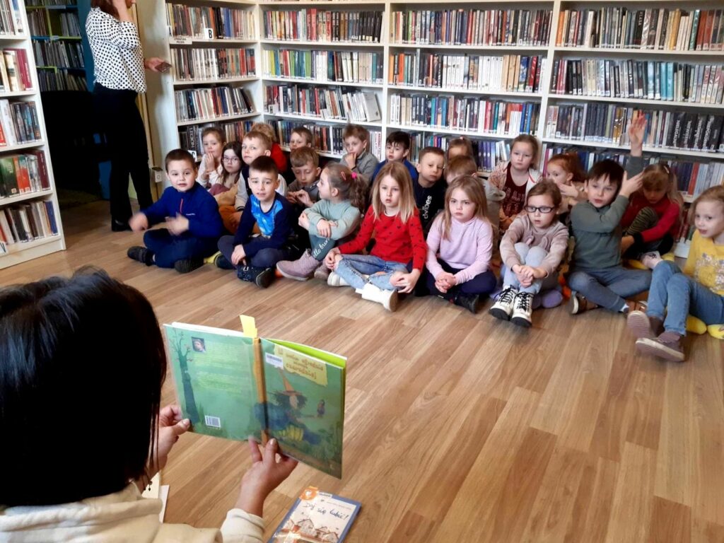 na zdjęciu widać dzieci siedzące w sali bibliotecznej i słuchające czytanej im książki
