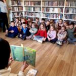 na zdjęciu widać dzieci siedzące w sali bibliotecznej i słuchające czytanej im książki