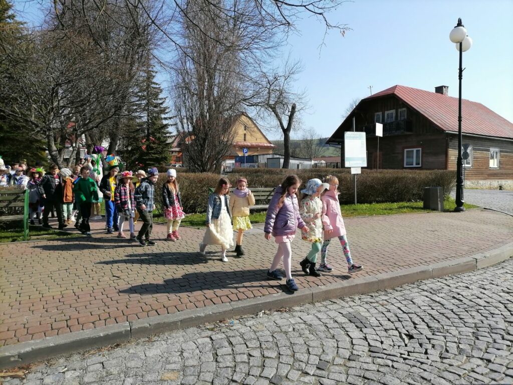 – Na zdjęciu widoczna jest grupa dzieci biorących udział w wiosennej imprezie plenerowej w Tyliczu i maszerujących przez tylicki rynek. Dzieci mają na sobie kolorowe wiosenne stroje. W tle drzewa i budynki.