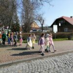 – Na zdjęciu widoczna jest grupa dzieci biorących udział w wiosennej imprezie plenerowej w Tyliczu i maszerujących przez tylicki rynek. Dzieci mają na sobie kolorowe wiosenne stroje. W tle drzewa i budynki.