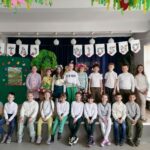 – Na zdjęciu widoczna jest grupa dzieci (10 chłopców i 9 dziewczynek) wraz z panią wychowawczynią pozujących do wspólnego zdjęcia pod napisem „Witaj wiosno”. Dzieci maja kolorowe wiosenne stroje. Jedna dziewczynka trzyma wykonany przez nich kwiat.