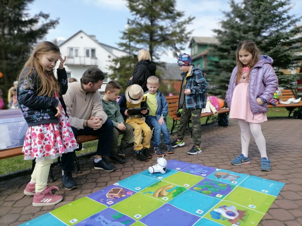 Na zdjęciu widoczna jest grupa dzieci obserwująca pokaz umiejętności edukacyjnego robota Photona prezentowanego przez pracownika biblioteki.