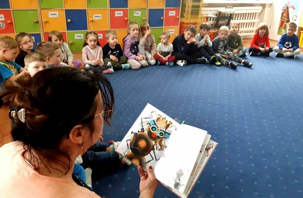 na zdjęciu widać dzieci siedzące na niebieskiej wykładzinie i czytającą im bibliotekarkę