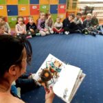 na zdjęciu widać dzieci siedzące na niebieskiej wykładzinie i czytającą im bibliotekarkę