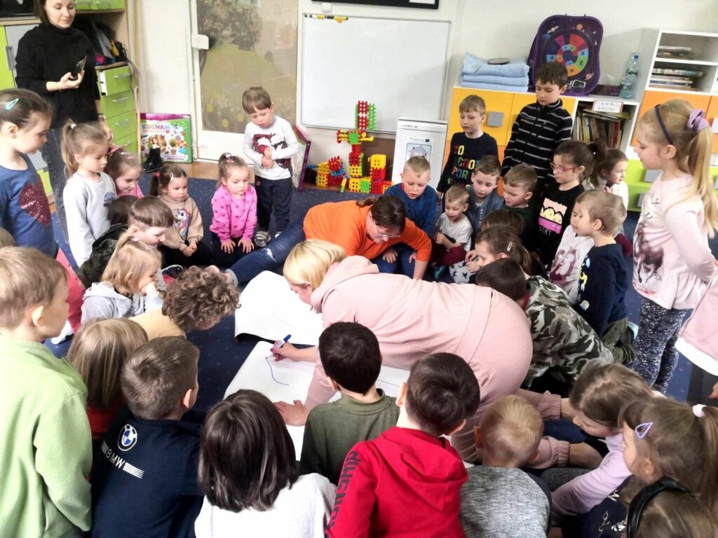 na zdjęciu widać dzieci w wieku przedszkolnym siedzące na podłodze wokół kolorowych ilustracji