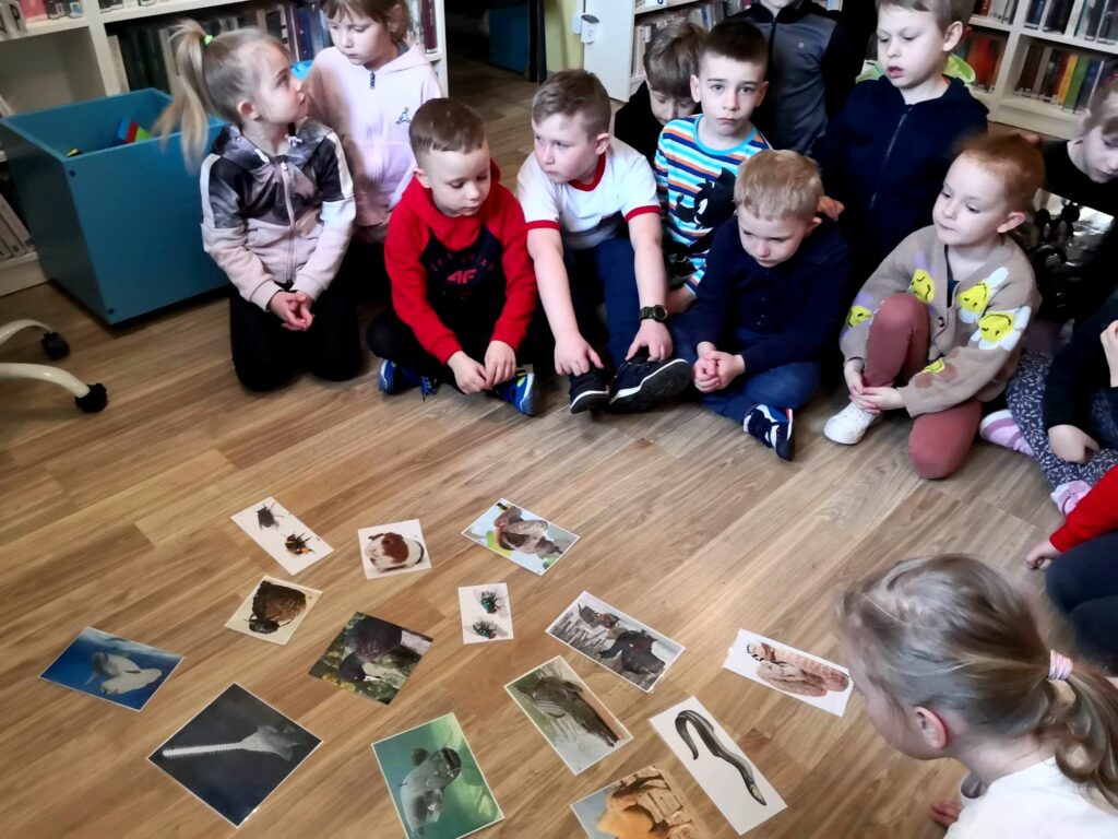 na zdjęciu widać dzieci siedzące na podłodze i przyglądające się fotografią przedstawiającym zwierzęta