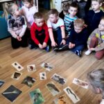 na zdjęciu widać dzieci siedzące na podłodze i przyglądające się fotografią przedstawiającym zwierzęta