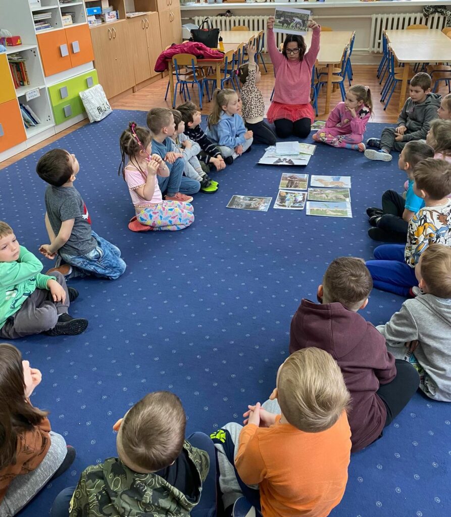 na zdjęciu widać dzieci w wieku przedszkolnym siedzące na niebieskim dywanie oraz bibliotekarkę pokazującą im ilustracje
