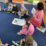 na zdjęciu widać dzieci w wieku przedszkolnym siedzące na niebieskim dywanie oraz bibliotekarkę pokazującą im ilustracje