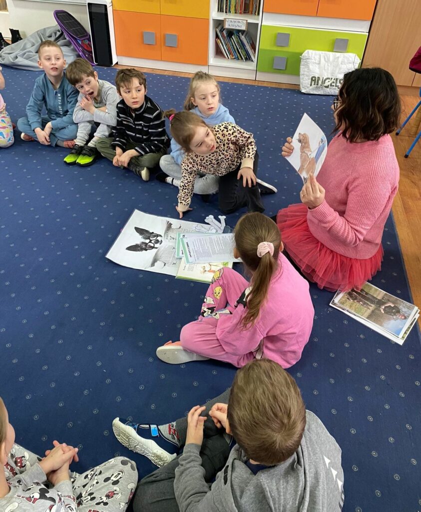 na zdjęciu widać dzieci w wieku przedszkolnym siedzące na niebieskim dywanie oraz bibliotekarkę pokazującą im ilustracje