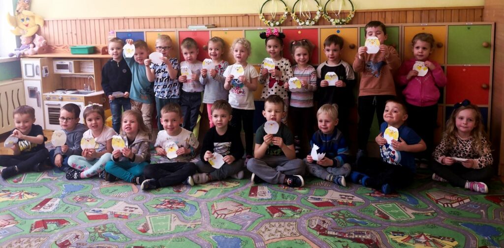 na zdjęciu widać stojące i siedzące dzieci w sali przedszkolnej trzymające papierowe kurczaki