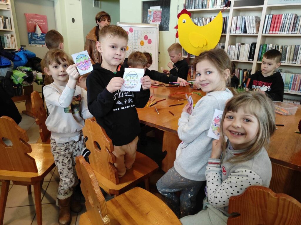 na zdjęciu widać salę biblioteczną, wokół stołu siedzą dzieci, wykonują pracę plastyczną