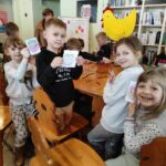 na zdjęciu widać salę biblioteczną, wokół stołu siedzą dzieci, wykonują pracę plastyczną