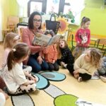 na zdjęciu widać salę przedszkolną, bibliotekarkę siedzącą na ziemi czytającą dzieciom książkę