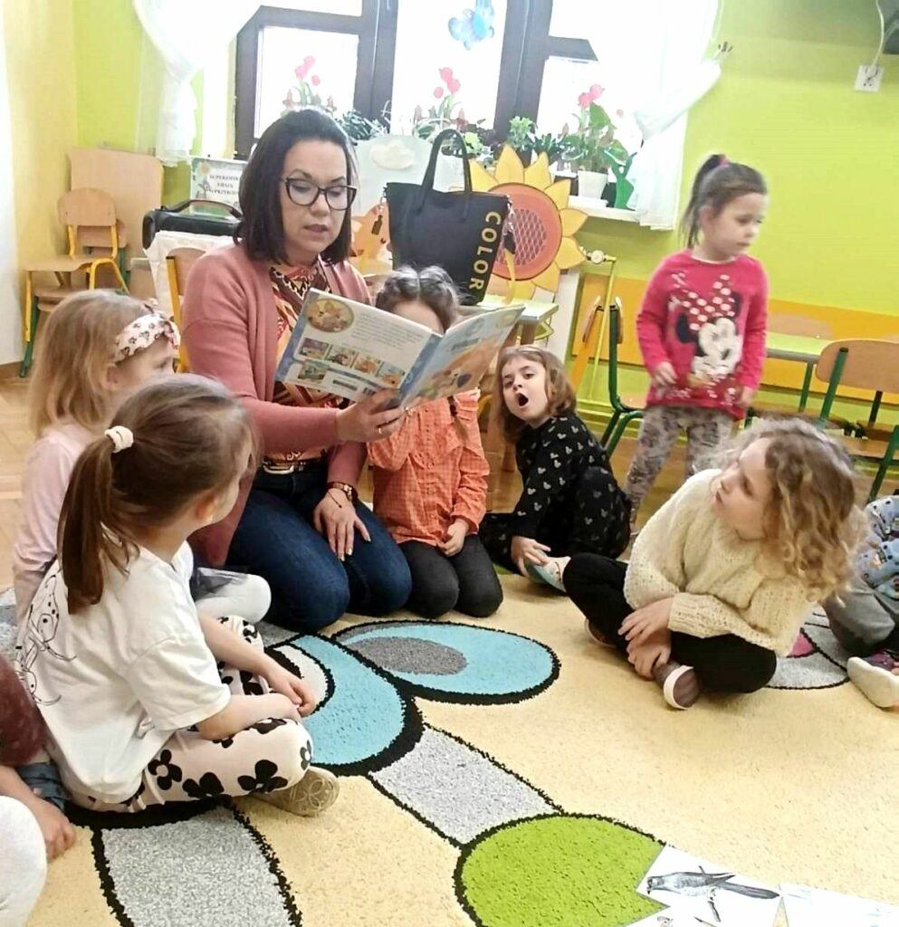 na zdjęciu widać salę przedszkolną, bibliotekarkę siedzącą na ziemi czytającą dzieciom książkę