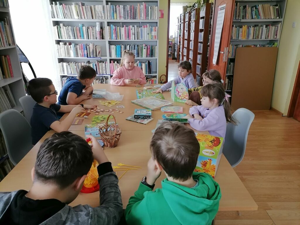 – Na zdjęciu widoczna jest grupa dzieci (4 chłopców i 4 dziewczynek), zapoznających się z książkami i grami związanymi z tematyką wielkanocną i wiosenną.