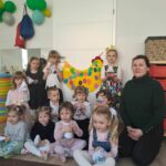 Na zdjęciu bibliotekarka i grupa 12 dzieci . Z tyłu dwie dziewczynki trzymaja żółtą kurkę z jajkami