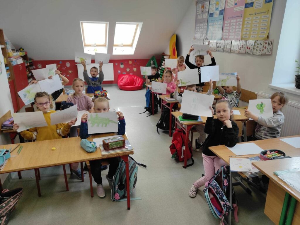na zdjęciu widać salę szkolną, przy stolikach siedzą dzieci prezentują swoje rysunki