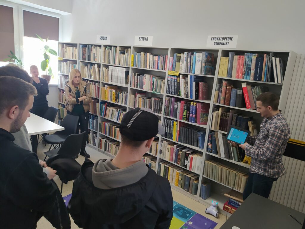 na zdjęciu sześc regałów na których ułożone sa ksiązki. Przed nimi grupa młodzieży z wychowawczynią oraz bibliotekarz przedstawiający photona