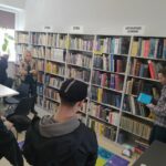 na zdjęciu sześc regałów na których ułożone sa ksiązki. Przed nimi grupa młodzieży z wychowawczynią oraz bibliotekarz przedstawiający photona