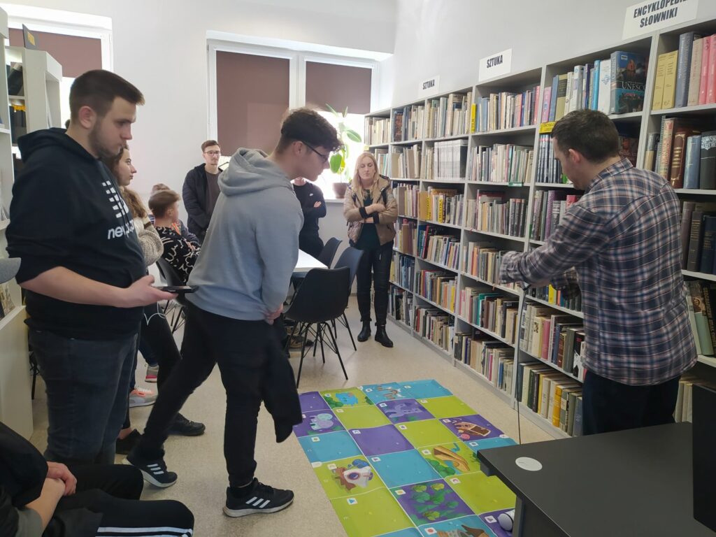 na zdjęciu sześc regałów na których ułożone sa ksiązki. Przed nimi grupa młodzieży z wychowawczynią oraz bibliotekarz przedstawiający photona