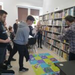 na zdjęciu sześc regałów na których ułożone sa ksiązki. Przed nimi grupa młodzieży z wychowawczynią oraz bibliotekarz przedstawiający photona