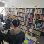 na zdjęciu sześc regałów na których ułożone sa ksiązki. Przed nimi grupa młodzieży z wychowawczynią oraz bibliotekarz przedstawiający photona