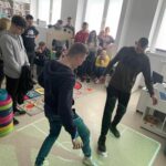 Na zdjęciu młodzież na tle regałów z książkami dla dzieci. Na pierwszym planie dwóch chłopców gra w piłkę nożną wyświetlana z fan flora