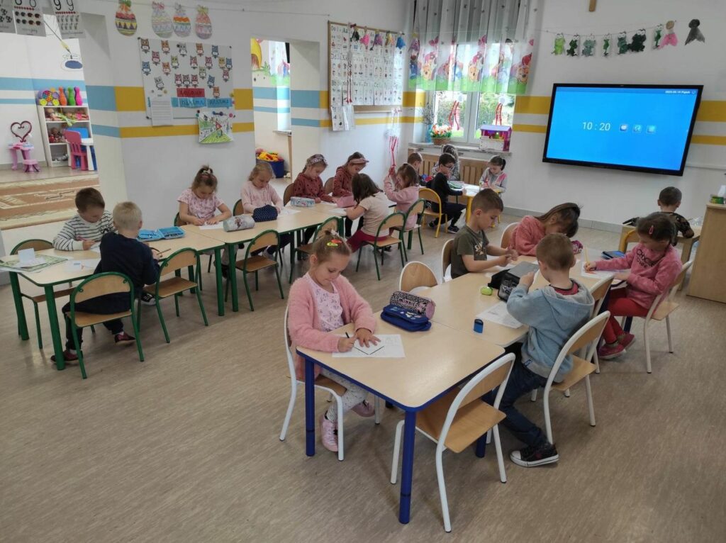 na zdjęciu widać salę przedszkolną, dzieci siedzą przy stolikach, wykonują pracę plastyczną