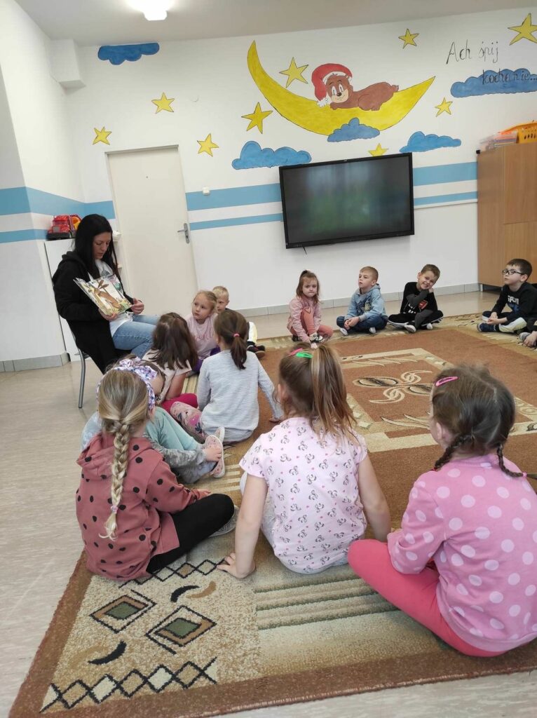 na zdjęciu widać salę przedszkolną, dzieci siedzą na dywanie, bibliotekarz czyta książkę