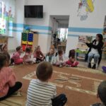 na zdjęciu widać salę przedszkolną, dzieci siedzą na dywanie, bibliotekarz czyta książkę