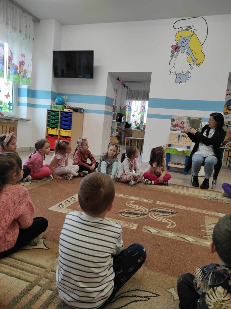 na zdjęciu widać salę przedszkolną, dzieci siedzą na dywanie, bibliotekarz czyta książkę