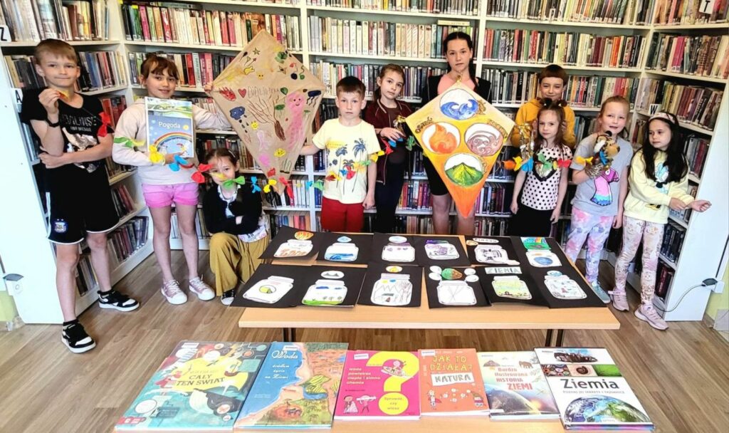 Na zdjęciu widać dzieci stojące w sali bibliotecznej na tle regałów. dzieci trzymają prace plastyczne