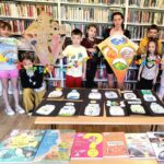 Na zdjęciu widać dzieci stojące w sali bibliotecznej na tle regałów. dzieci trzymają prace plastyczne